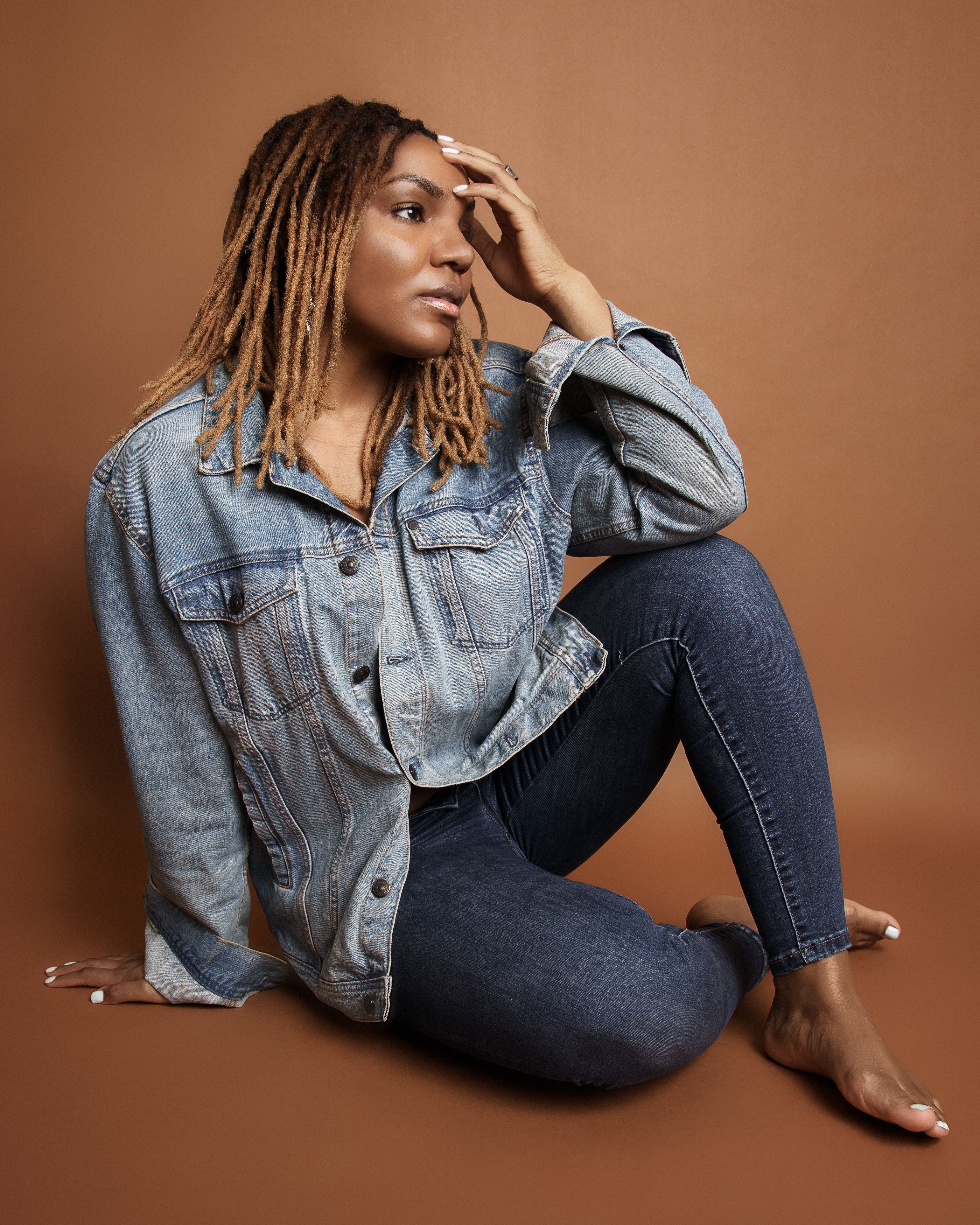 Relaxed woman in denim jacket and jeans posing barefoot in studio setting, exuding casual style.
