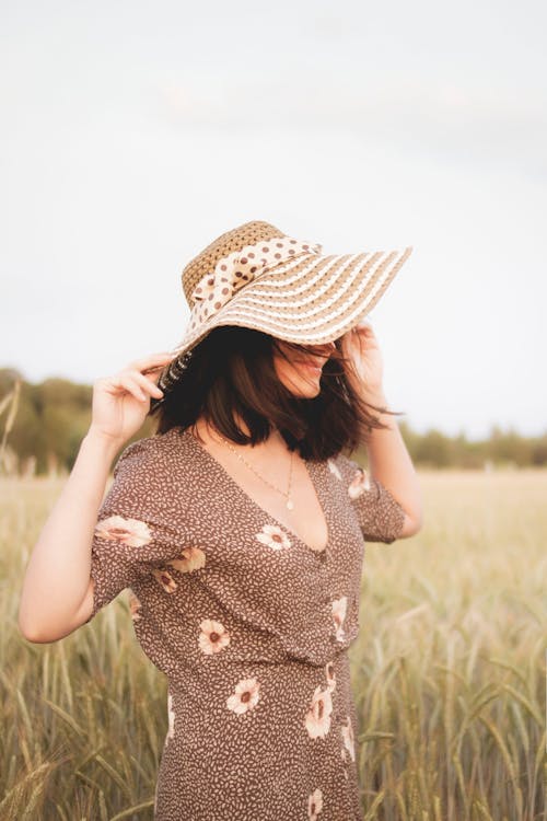 Free Side View Photo of Woman in Floral Dress and Sun Hat Standing in Grass Field Stock Photo