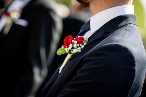 Free Close-up of a stylish man in a suit adorned with a vibrant rose boutonniere at a daytime wedding. Stock Photo