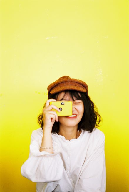 Happy woman in white top taking photo against bright yellow backdrop.