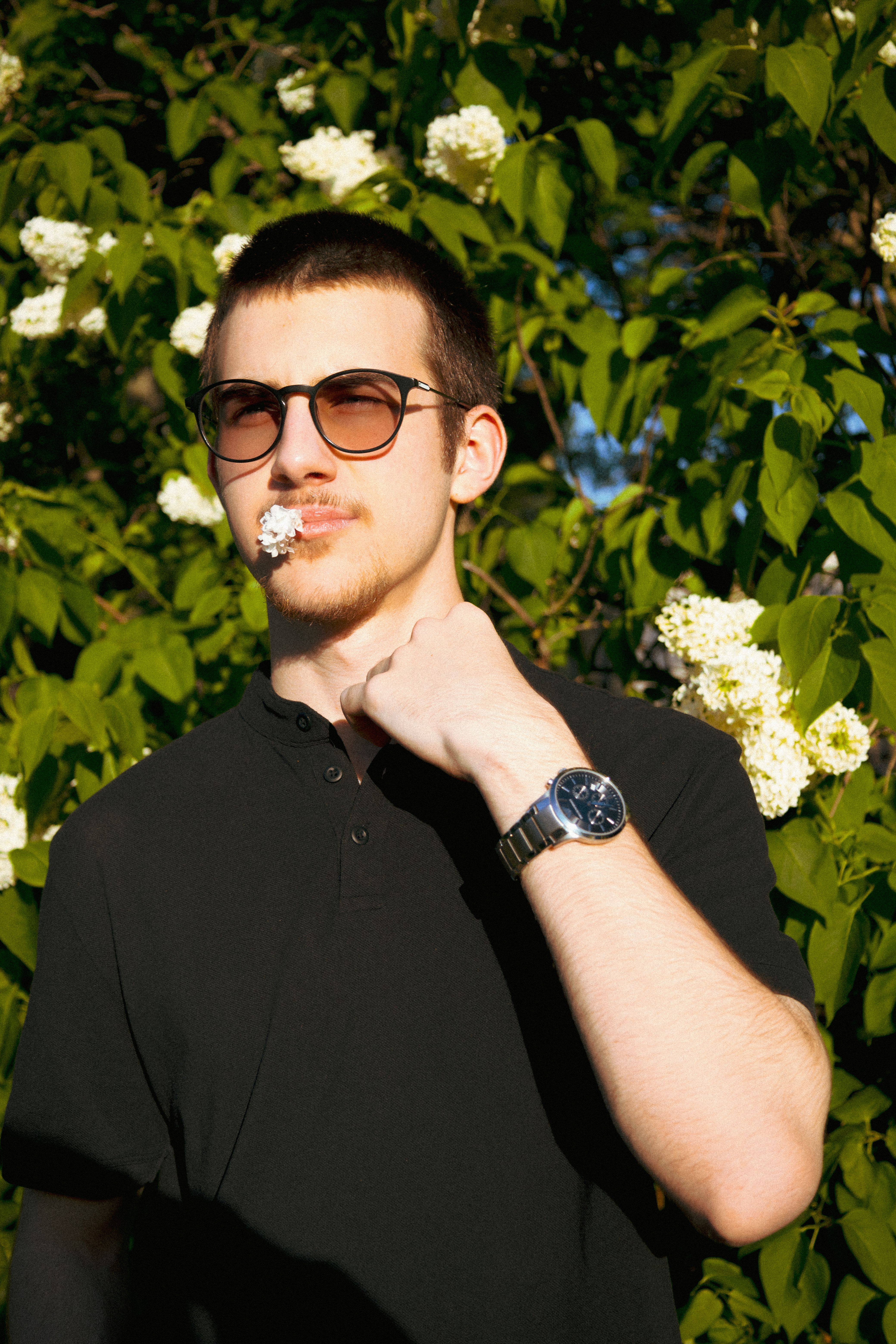 Young man posing with sunglasses and flowers in a garden, enjoying the sunlight and greenery.