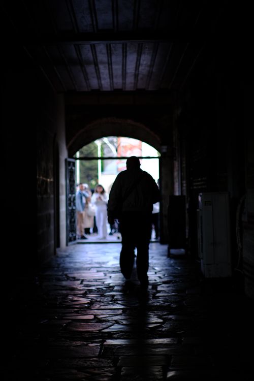 Free A silhouette captures a man walking through a dimly lit passageway with a group visible at the entrance, creating a moody urban scene. Stock Photo