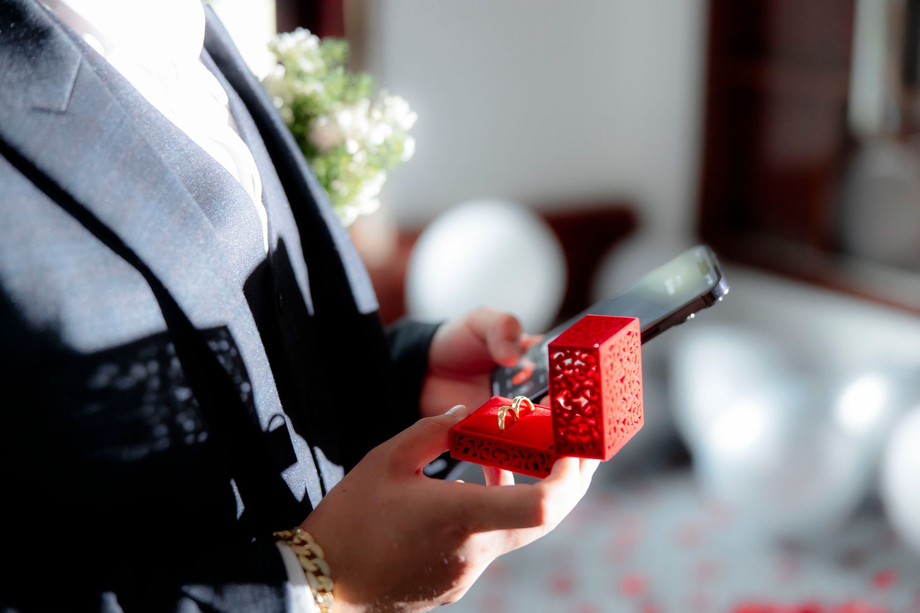 A groom in formal attire holds a red ring box and smartphone during a wedding ceremony.