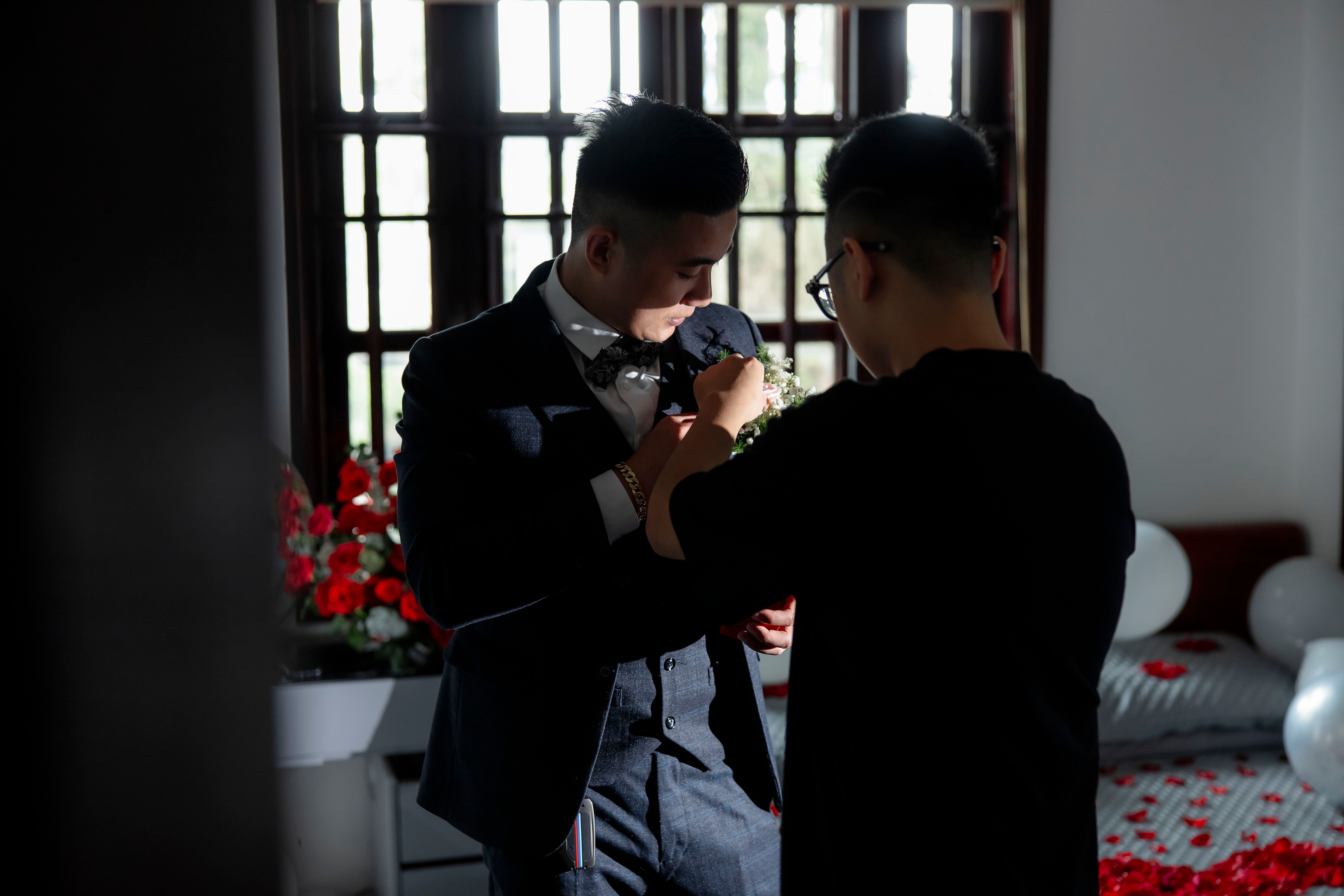 Groom in elegant suit having boutonniere adjusted before wedding ceremony, indoors setting.