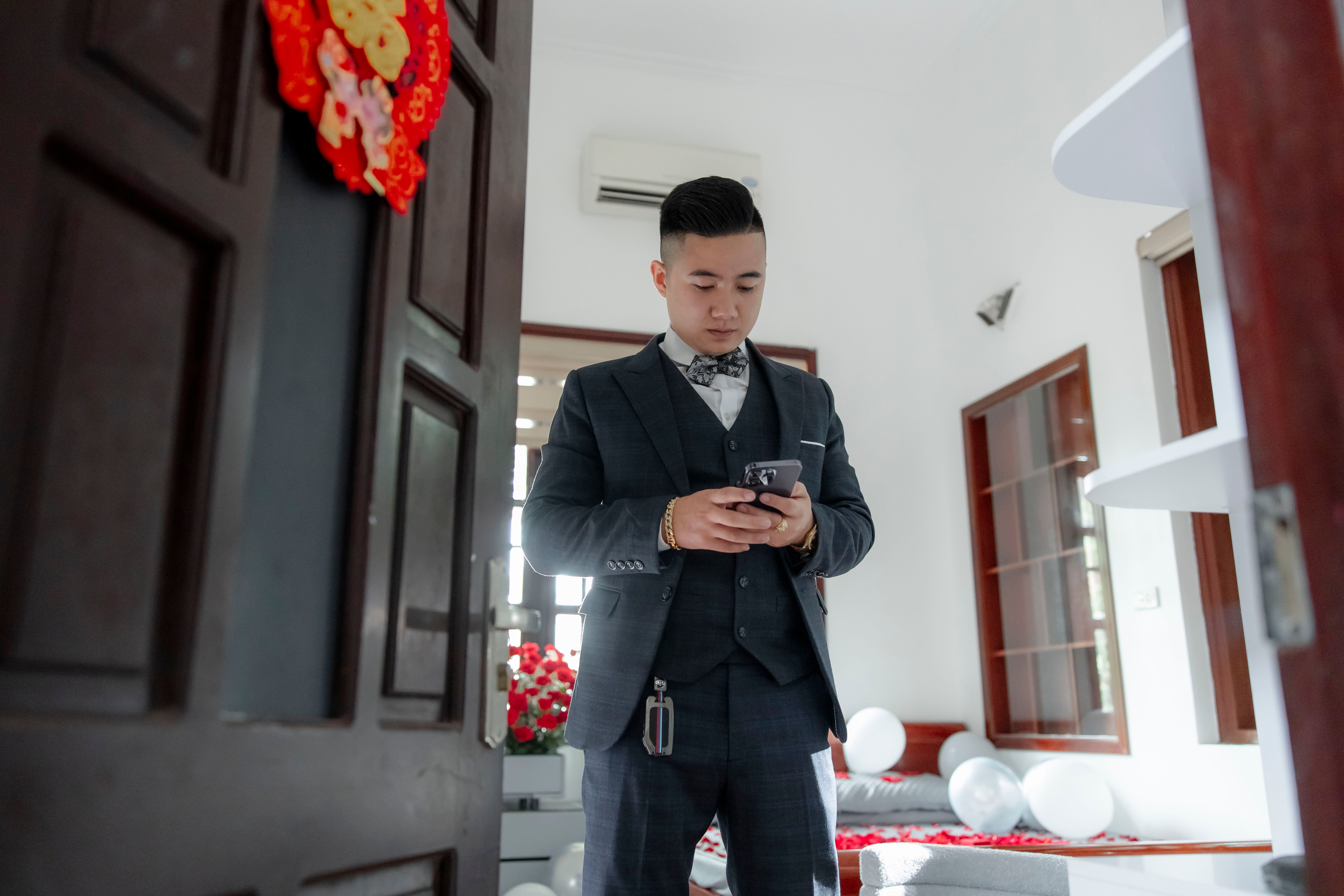 A groom in a suit checks his smartphone indoors, preparing for a wedding ceremony.