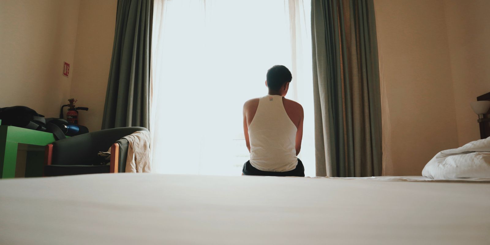 A man sits indoors facing a bright window, lost in thought, casting an introspective mood.