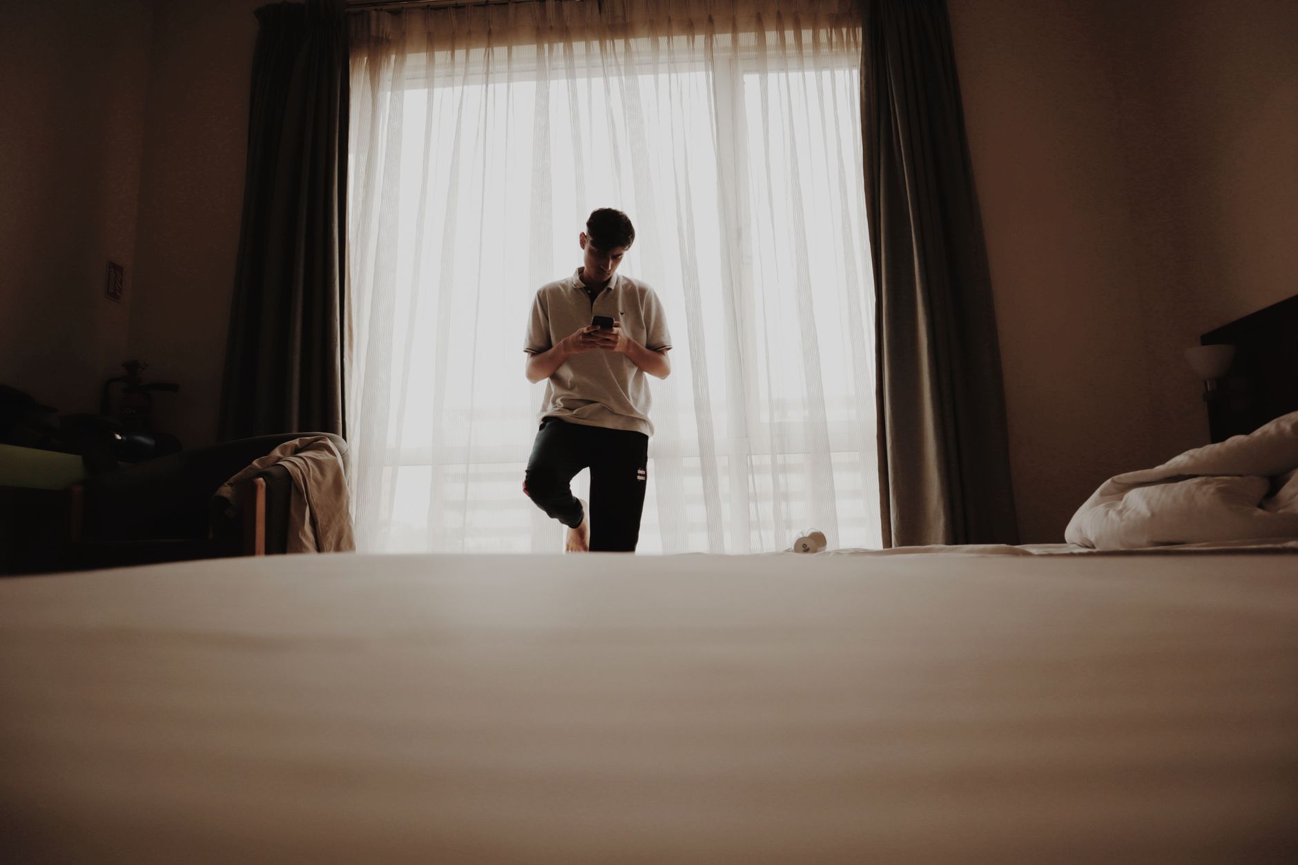 A young man stands in a modern bedroom looking at his phone — the suspended animation of a life waiting to begin.