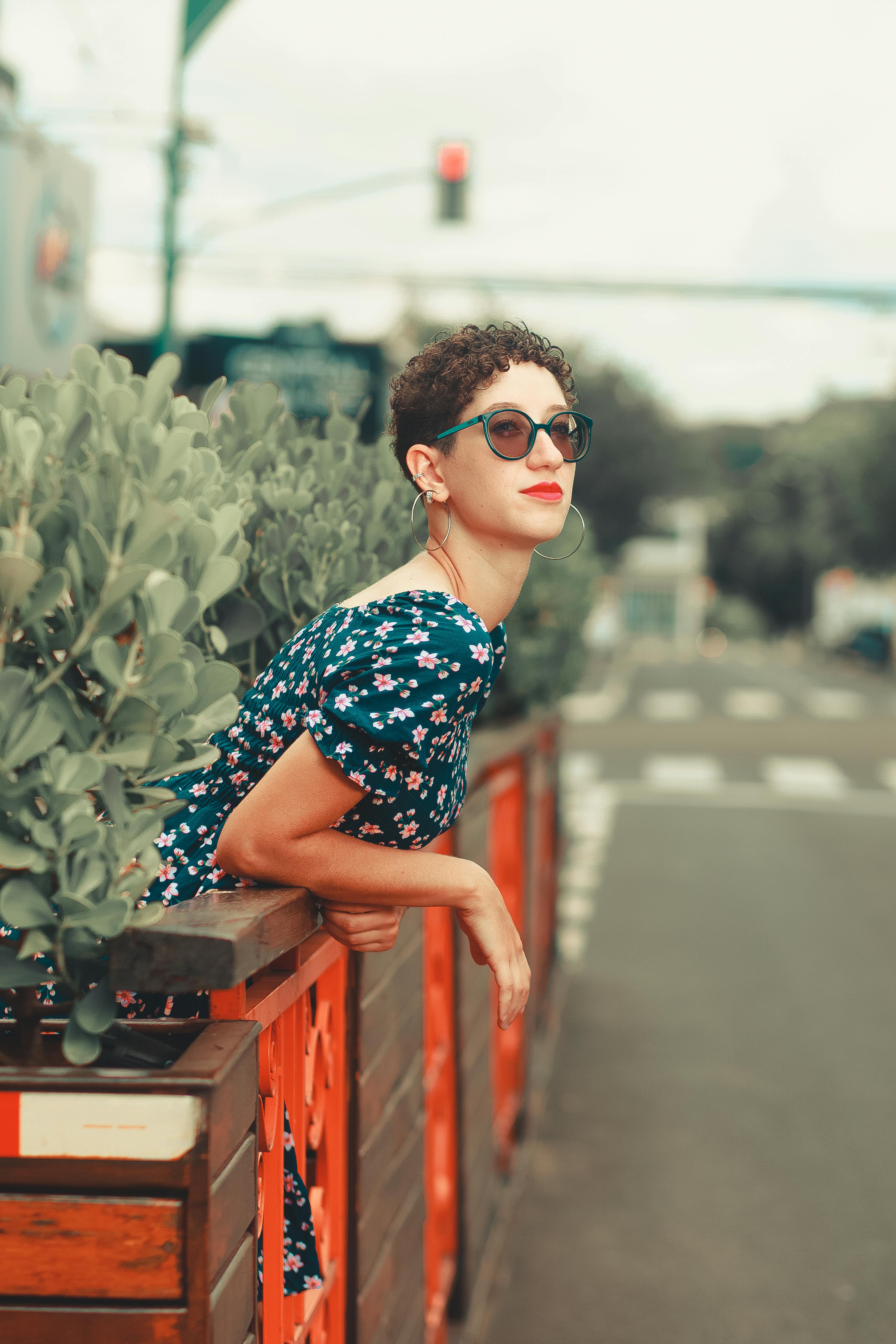 Young woman leans on urban patio railing in summer, wearing sunglasses and floral dress.
