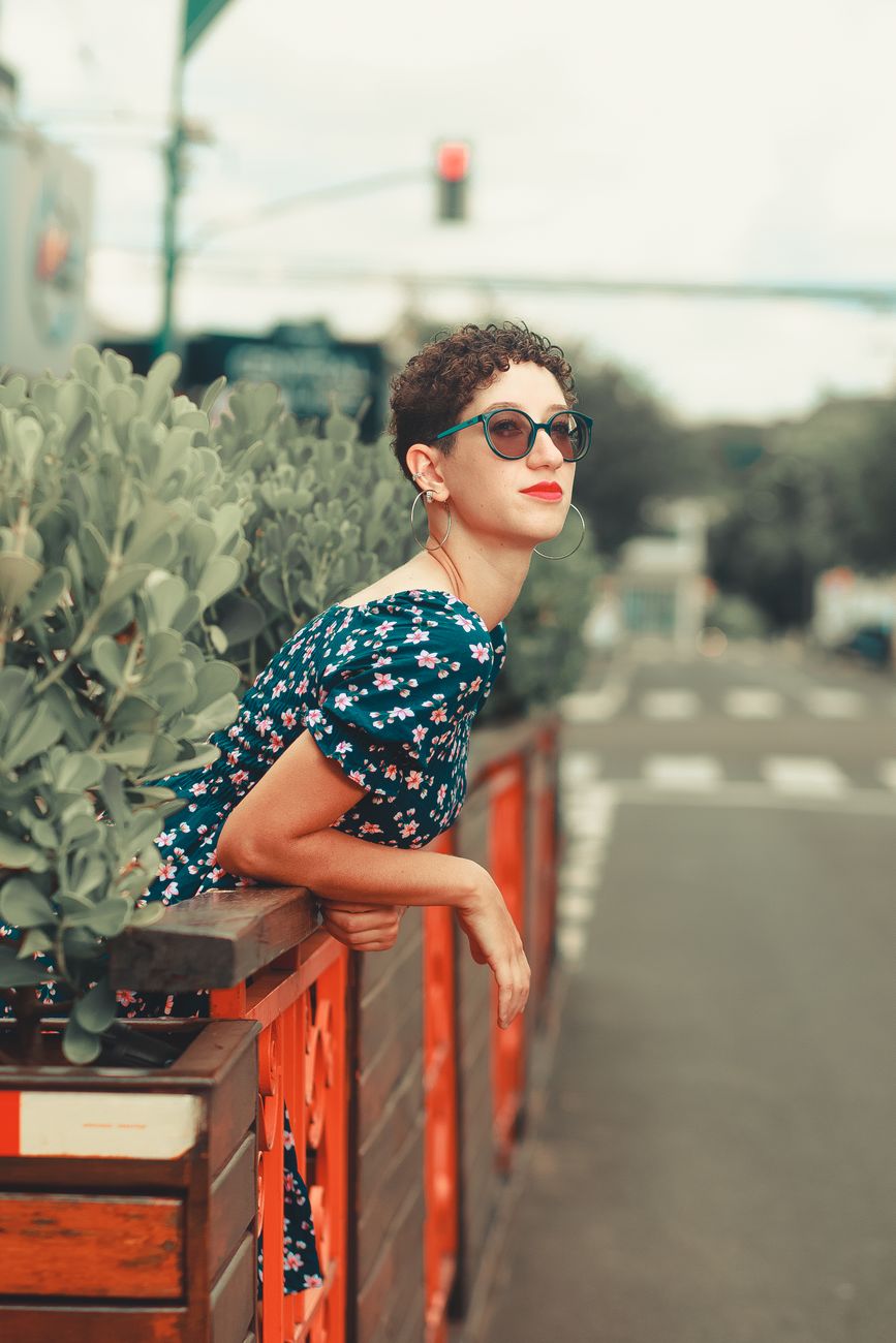 Young woman leans on urban patio railing in summer, wearing sunglasses and floral dress.