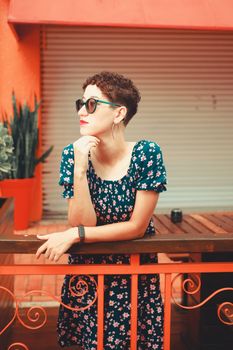 Portrait of a woman in a floral sundress wearing sunglasses, leaning on an urban railing.