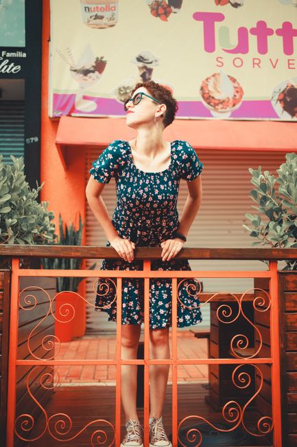 Woman in a floral dress standing by a café railing on a sunny day.