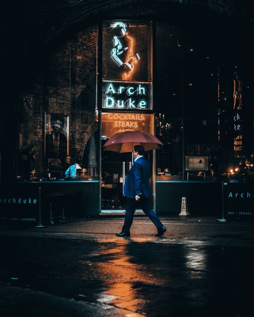 A man in a blue suit walks past the Arch Duke in London, holding a red umbrella on a rainy night.