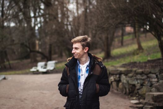 Cheerful young man in a jacket smiling with hands in pockets, outdoors in a park.