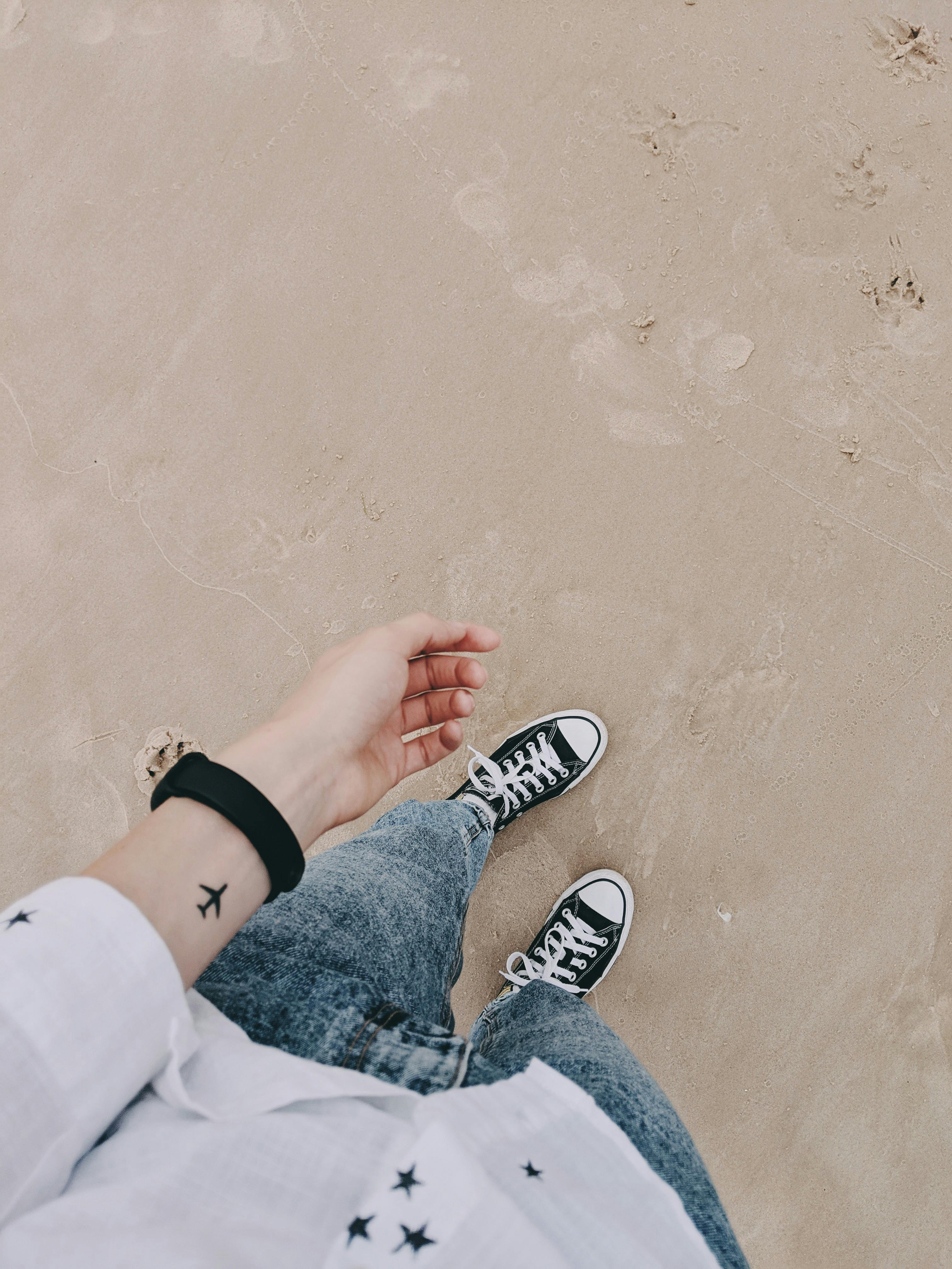 A person with a tattoo enjoys a casual beach walk wearing denim jeans and sneakers on sandy shore.