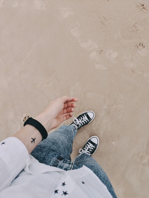A person with a tattoo enjoys a casual beach walk wearing denim jeans and sneakers on sandy shore.