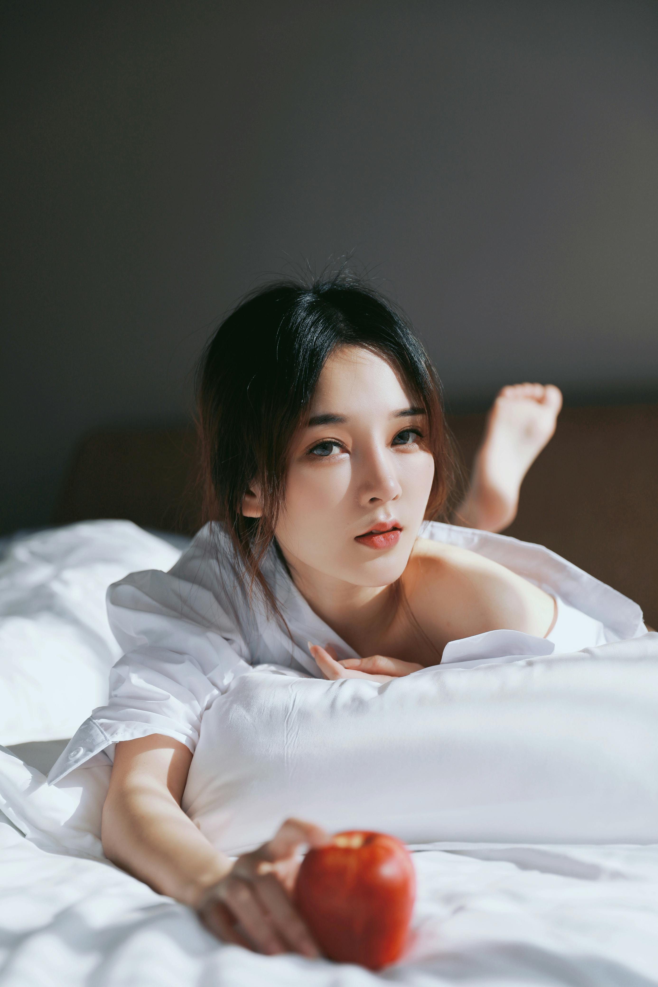 A serene portrait of a young woman lying on a bed, holding a red apple, evoking a sense of tranquility.