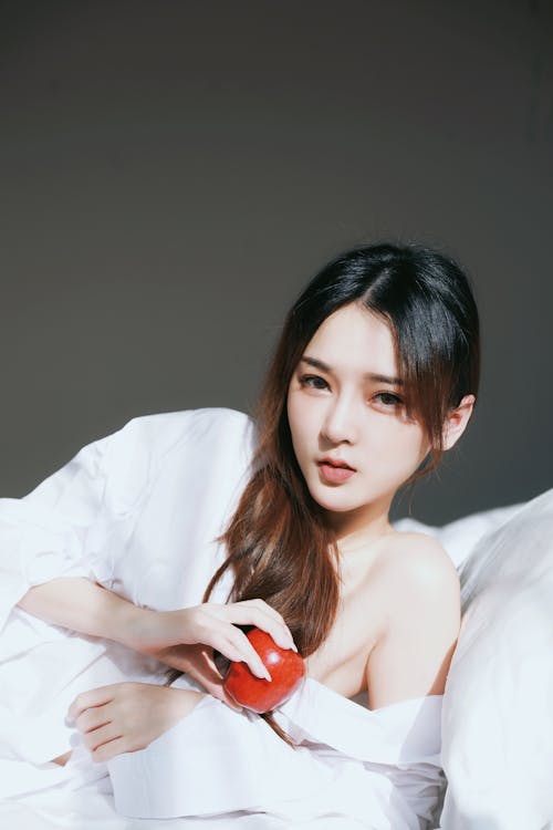 A woman in white shirt laying in bed with red apple