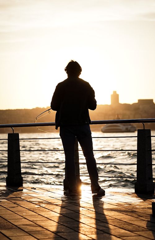 Free A man fishing at sunset with a city skyline backdrop, creating a serene silhouette scene. Stock Photo