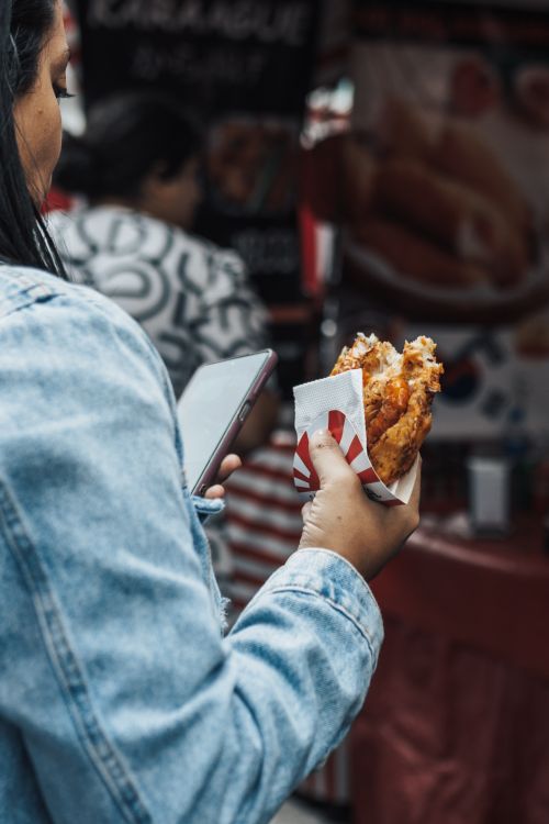 Free A woman holds a wrapped snack at a bustling outdoor food market, using her phone. Stock Photo