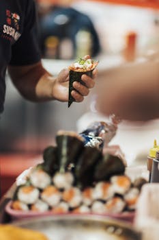Close-up of a chef serving fresh temaki sushi at a bustling food stall with stacks of rolls.