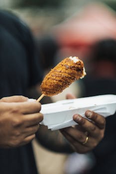 Man holding a crispy fried corn dog in a paper tray on a street. Perfect for food festival and street food promotions.