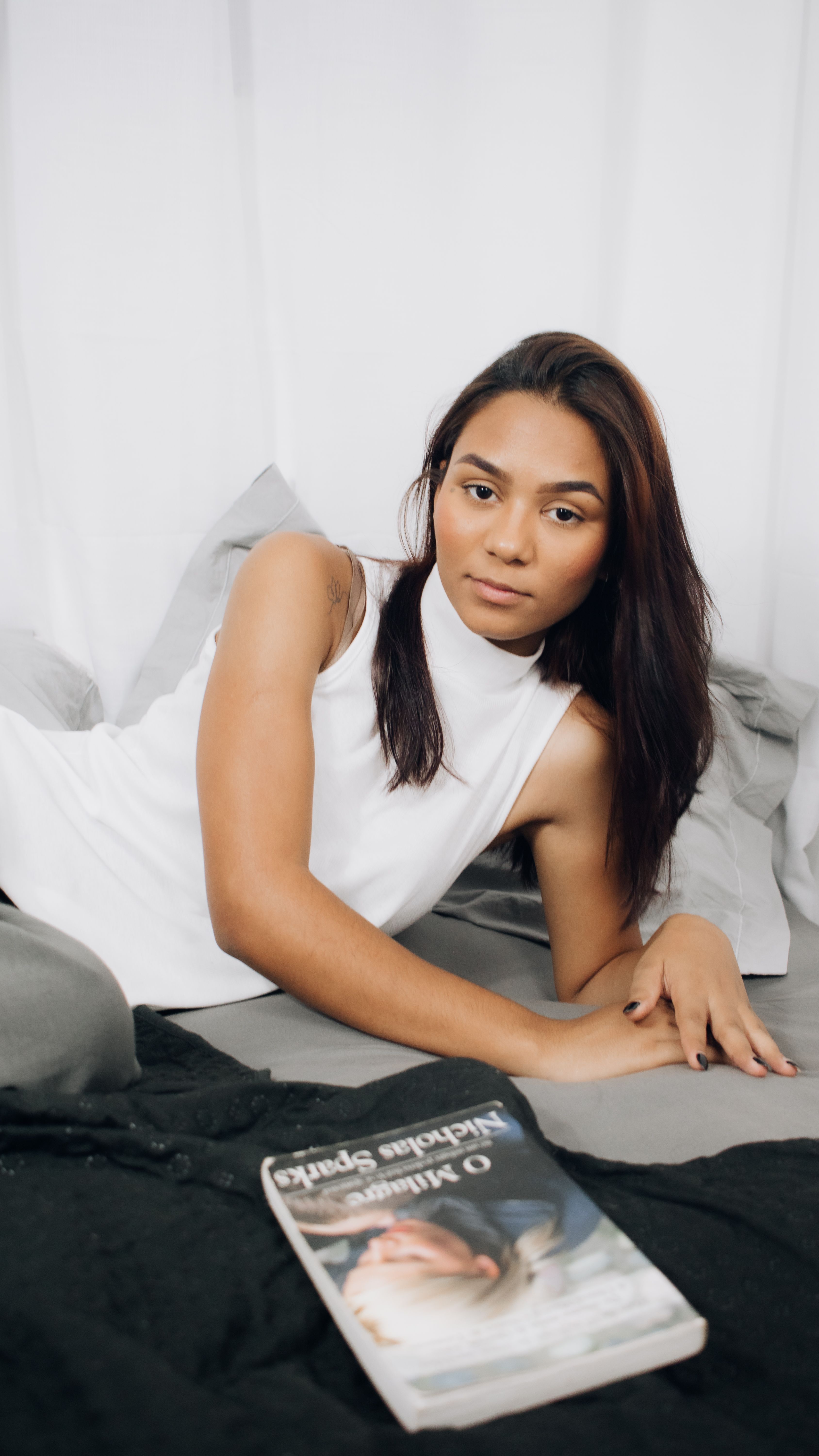 A woman in a white top lies on a bed, engaging in relaxation and reading.