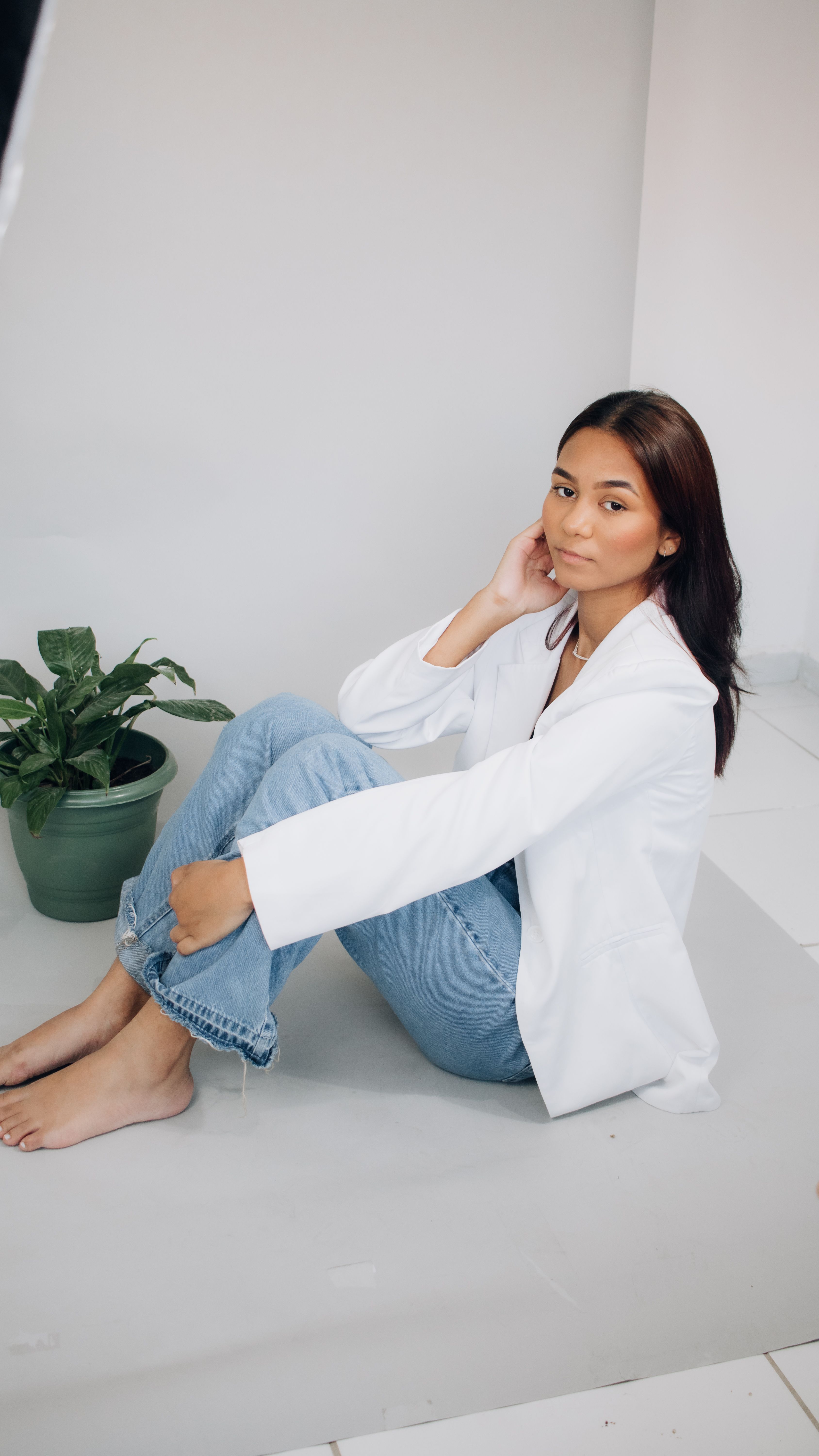 A young woman sits with poise in jeans and a white jacket, embodying relaxed elegance.