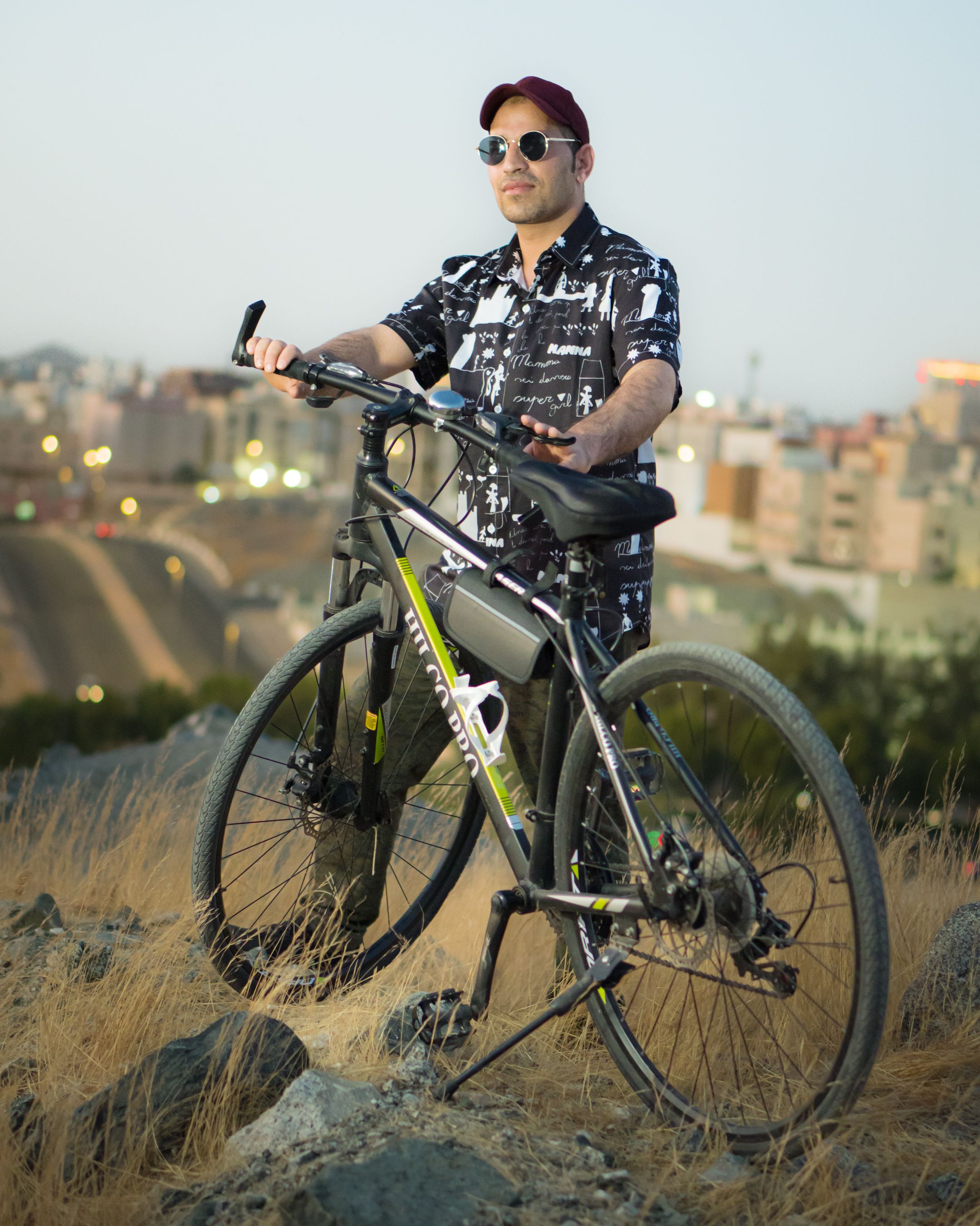 Adult man in sunglasses stands with mountain bike on a hill overlooking Makkah city at dusk.