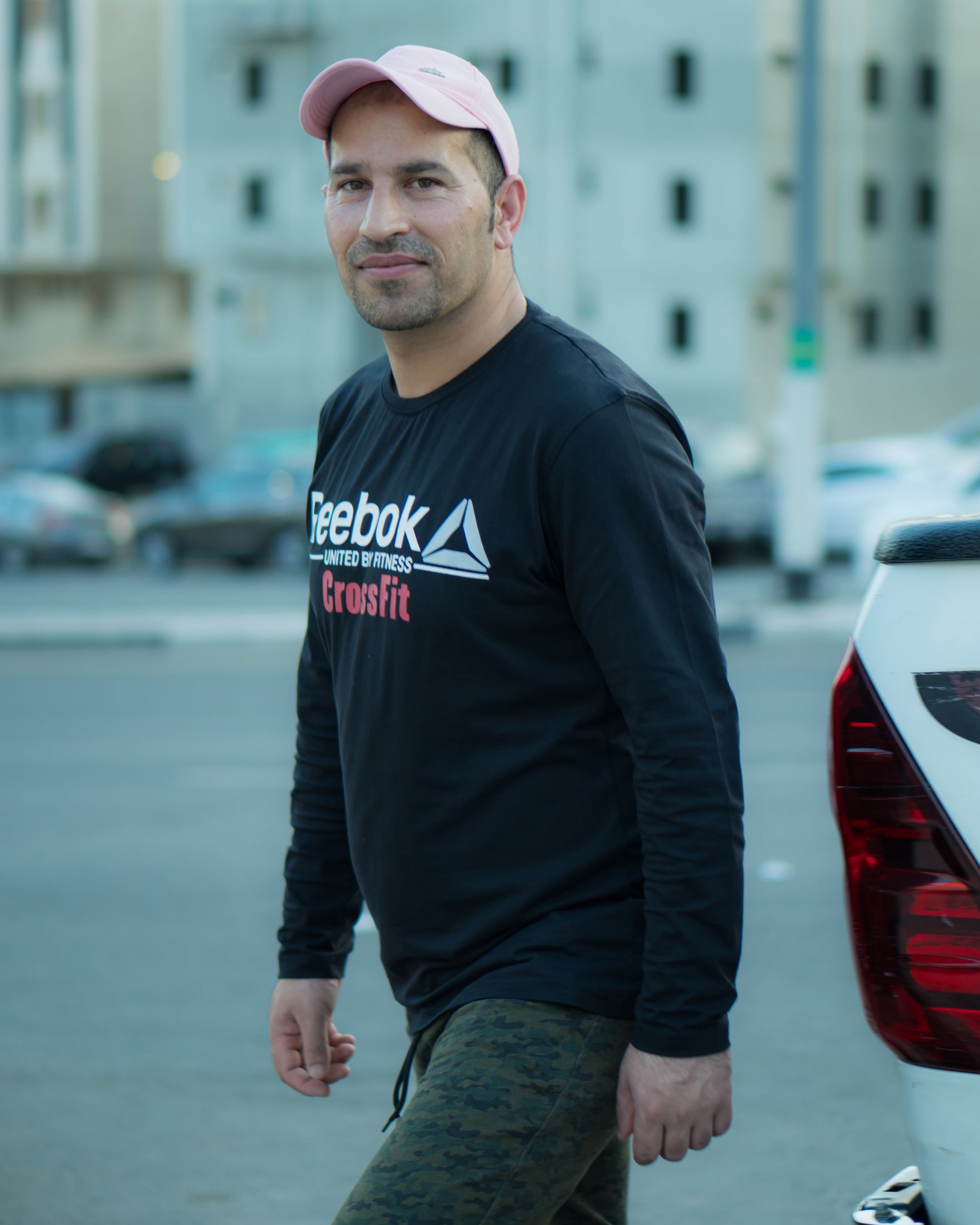 Man wearing black sweatshirt and pink cap standing outdoors, urban background.