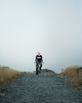 A man rides a bicycle along a solitary gravel path surrounded by dry grass, evoking a sense of adventure.