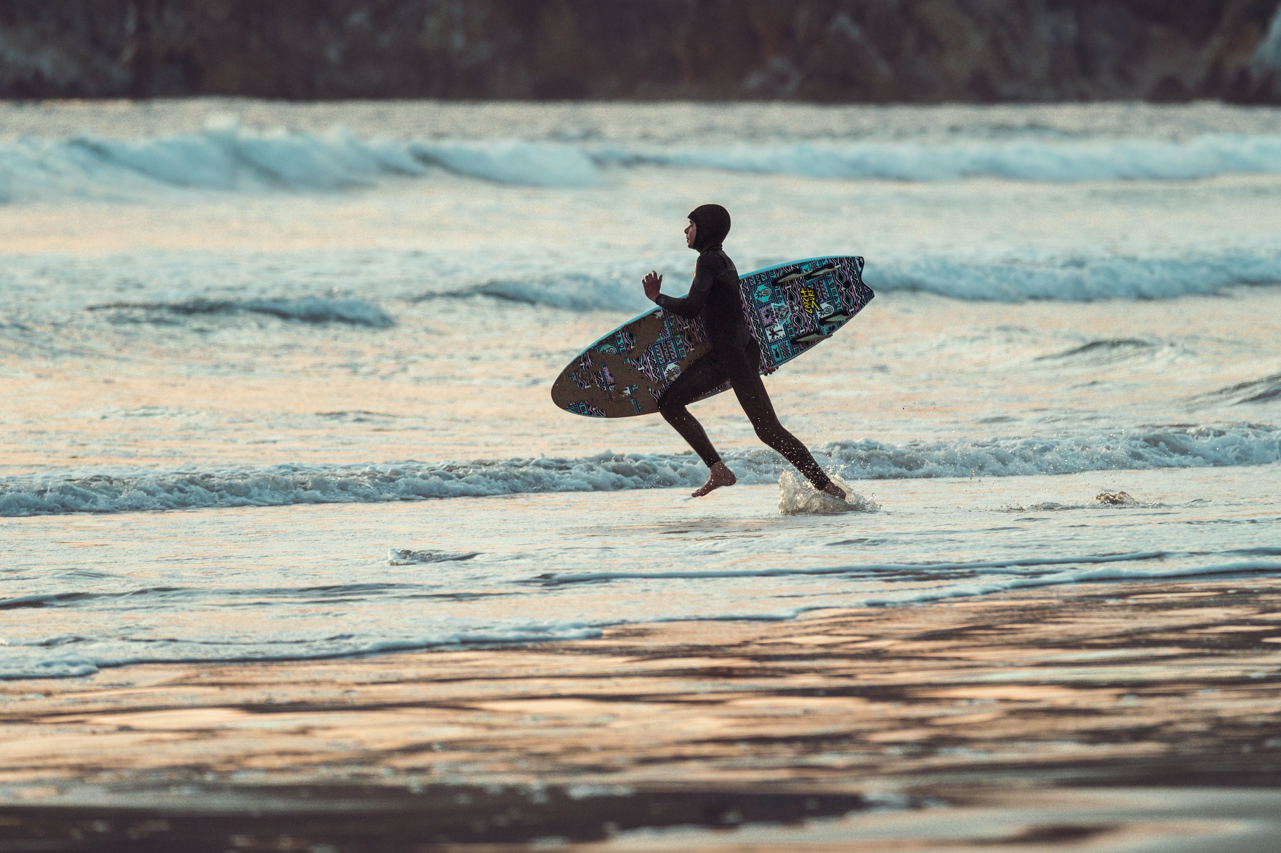 A surfer in a wetsuit runs along Pismo Beach carrying a surfboard during sunset.