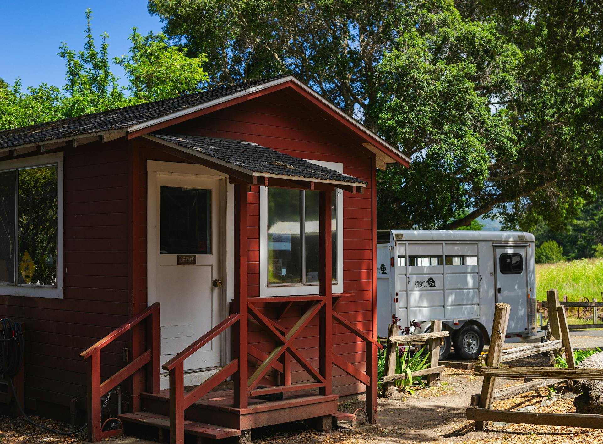 A small red cabin with a trailer surrounded by trees in a rural area under a bright sky.