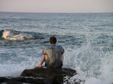 A person sits on rocks by a wavy ocean, capturing a serene moment of contemplation and natural beauty.
