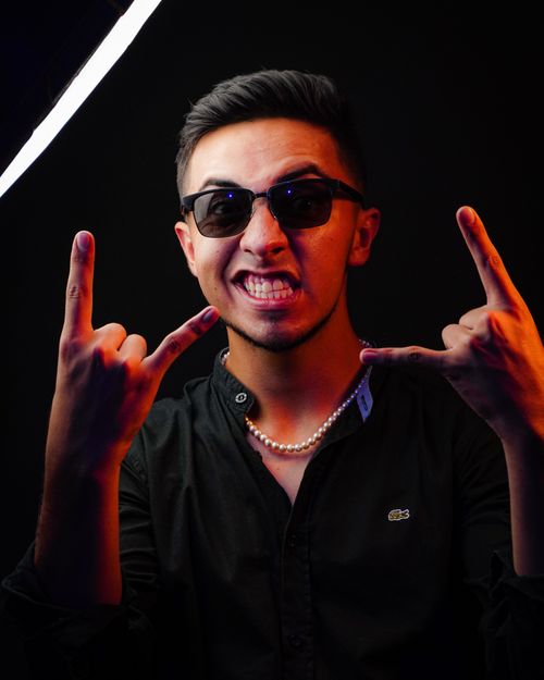 Free Portrait of a confident man wearing sunglasses, making rock sign gesture in a dark studio setting. Stock Photo