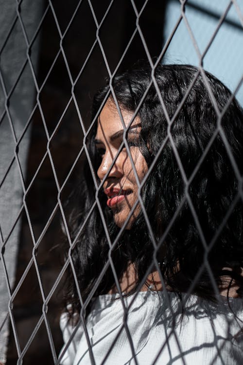 Free Close-up Portrait Photo of Smiling Woman Standing Behind Chain-link Fence Looking into the Distance Stock Photo