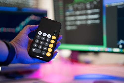 Free A person uses a smartphone calculator app in a modern office with computer screens displaying charts. Stock Photo