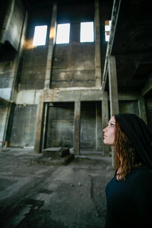 Foto De Vista Lateral De La Mujer En Vestido Negro Mirando Hacia Arriba Mientras Está De Pie En Un Edificio Abandonado