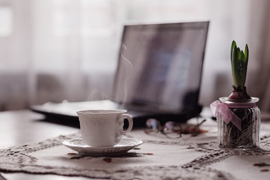 A serene indoor workspace featuring a steaming teacup, laptop, and potted spring bulb, all on a decorative tablecloth.