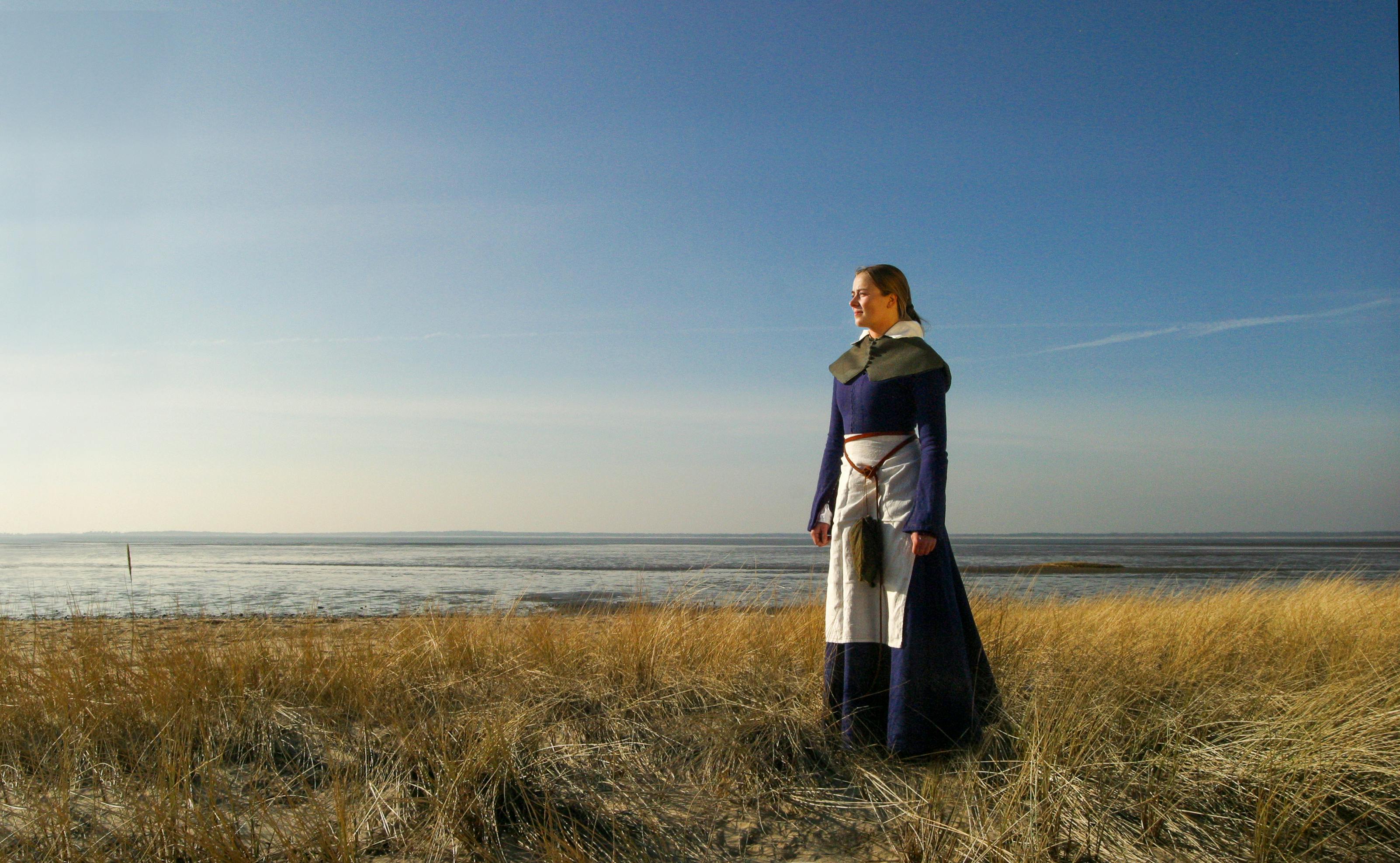 Woman in medieval attire standing on a coastal field during the day