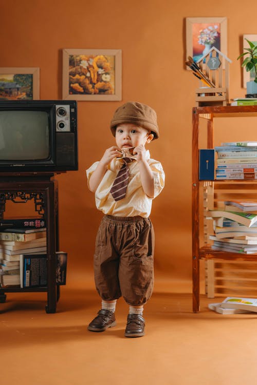 A small boy in a hat and brown pants standing in front of a television