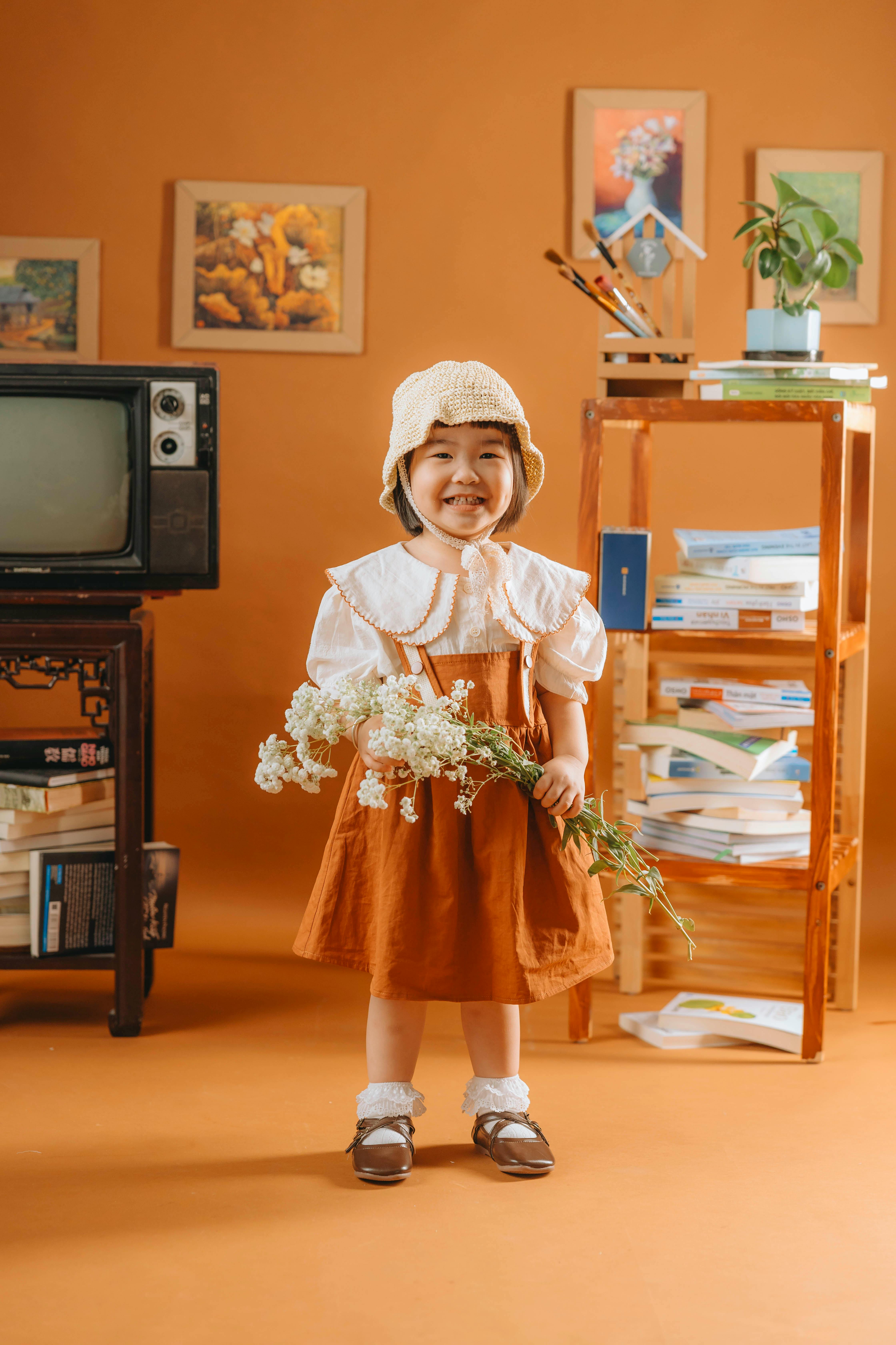 Smiling child in vintage outfit holding flowers in a cozy indoor setting.