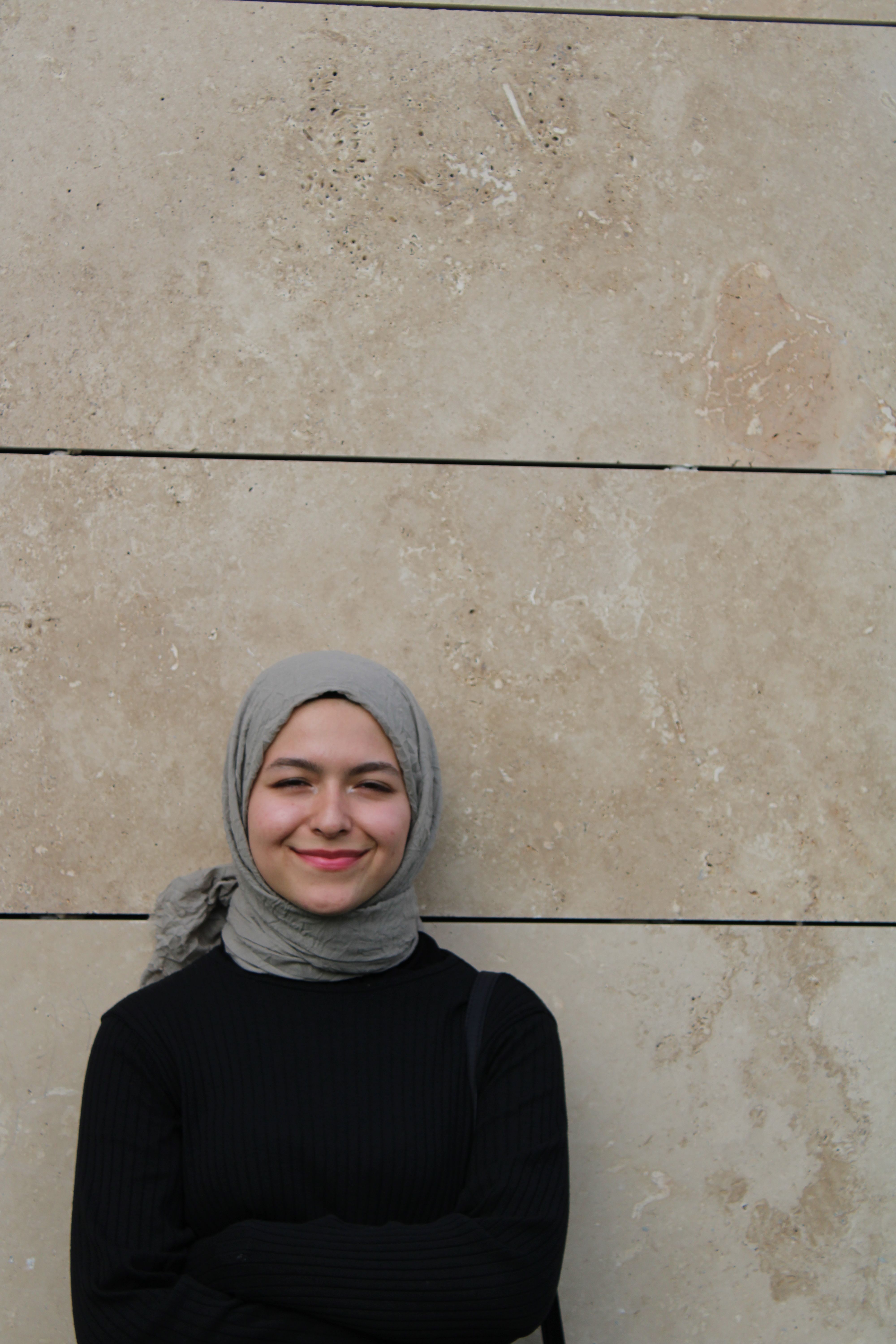 Portrait of a young woman in a black gown and hijab smiling against a stone wall.