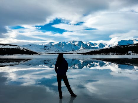 在阿根廷巴塔哥尼亚，一名女子站在冰冻的湖面上，湖面倒映着白雪皑皑的山脉