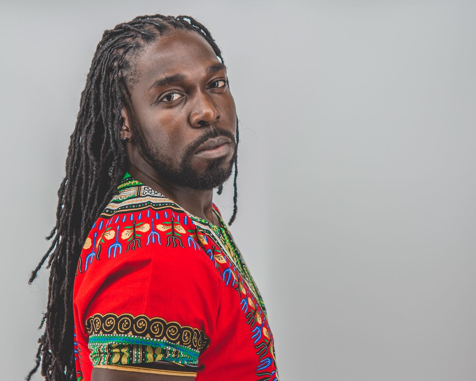 A stylish man with dreadlocks in vibrant African clothing against a neutral background, exuding confidence and cultural pride.
