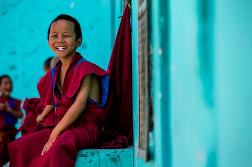 Free A young monk smiling joyfully while sitting outdoors in a vibrant red robe, capturing cultural essence. Stock Photo