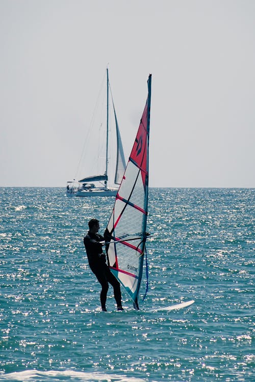 A man windsurfing in the ocean with a sailboat in the background