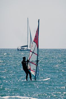 A man enjoys windsurfing on the open ocean with a sailboat in the background.