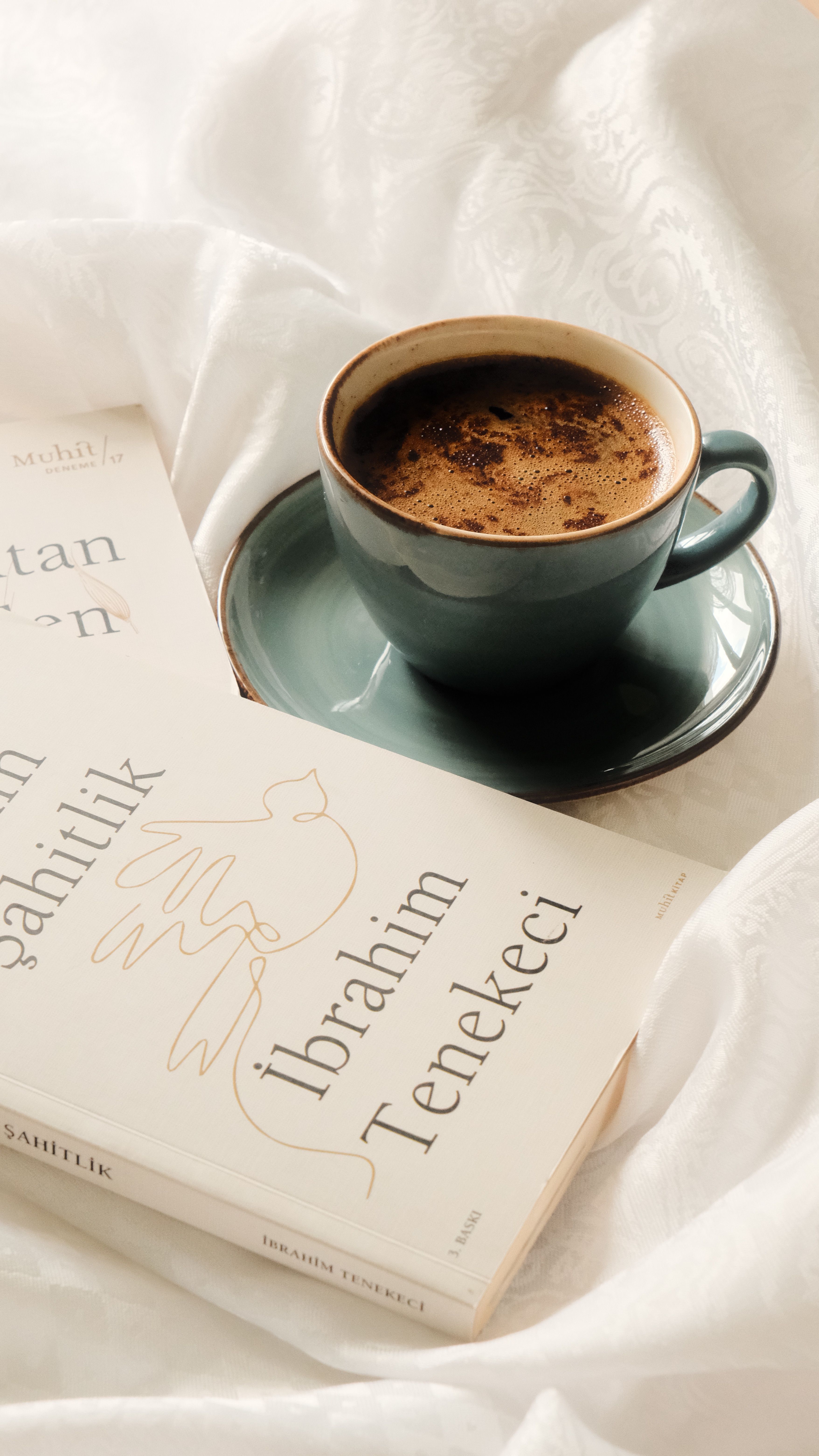 A cup of coffee and a book on a soft white sheet create a cozy reading nook.