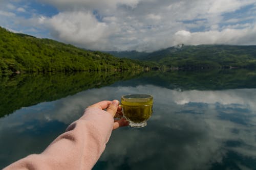 A hand holding a cup of matcha tea over a gorgeous view of the lake & mountains reflecting in the water