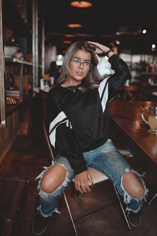 Foto Fokus Selektif Wanita Dengan Sweatshirt Hitam Putih Dan Jeans Distressed Biru Duduk Di Kursi Lipat Baja Di Sebelah Meja Kayu Di Kafe